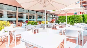 An outdoor dining area with white tables and chairs, large umbrellas, and a building with glass windows in the background. - BLUESEA Montevista Hawai (Lloret de Mar)