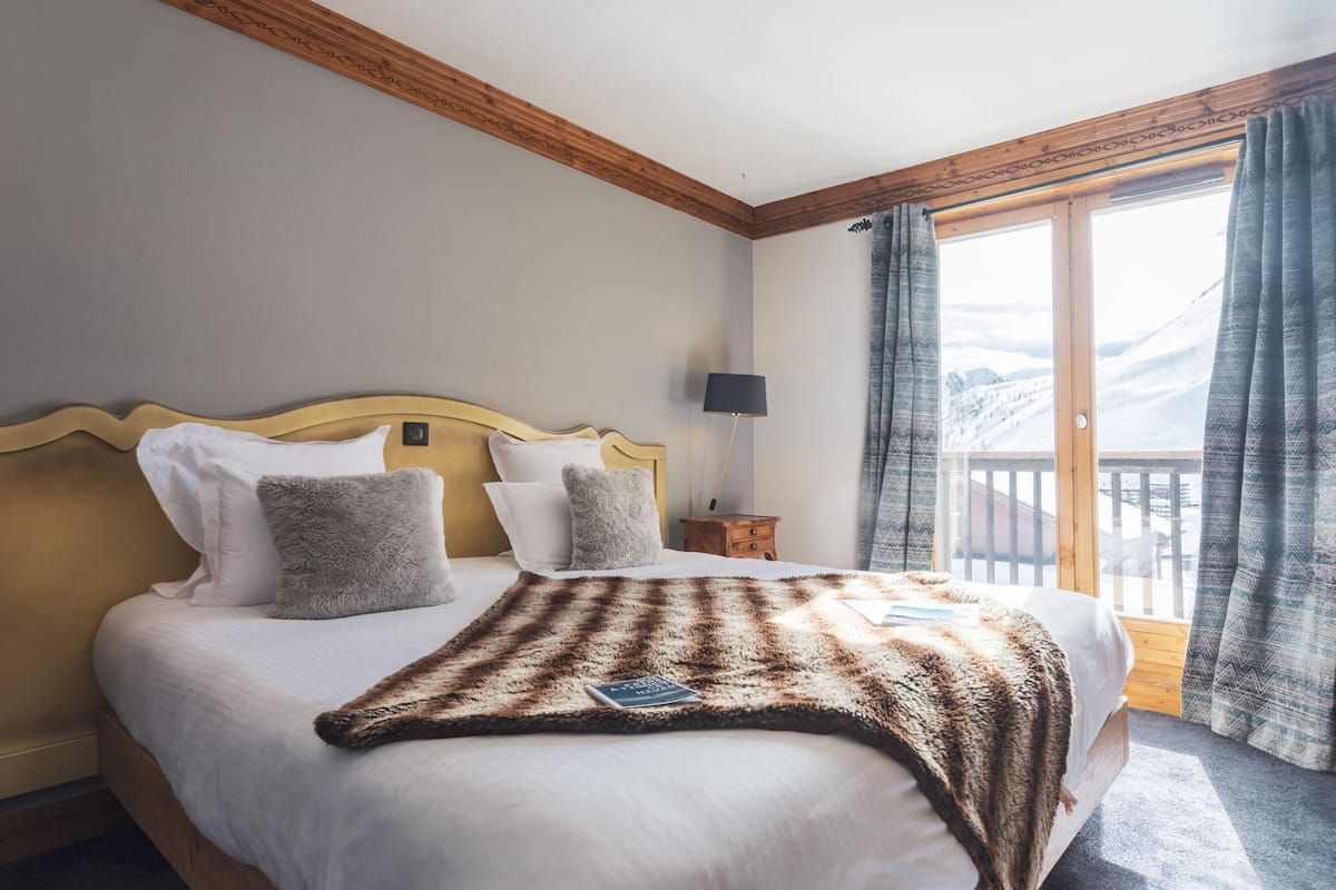 Une chambre avec une tête de lit en bois, un grand lit avec une literie blanche, une couverture douce et moelleuse, une table de chevet avec une lampe, et une vue sur les montagnes depuis un balcon.
