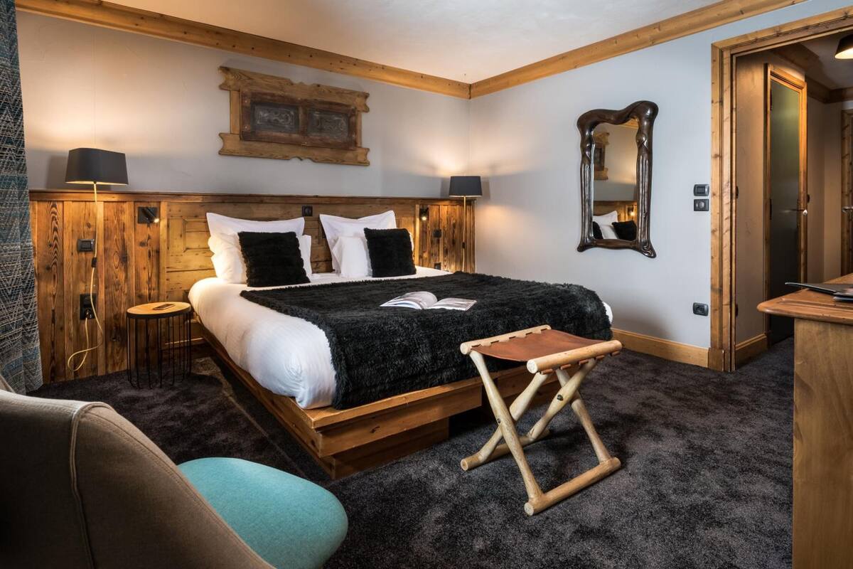 Une chambre à coucher avec un lit en bois, un petit tabouret en bois, une table de chevet et un miroir accroché au mur.