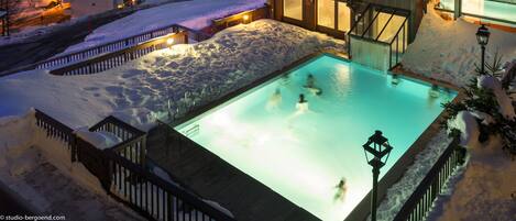 A snow-covered mountain resort at night with illuminated buildings, a swimming pool, and ski lifts in the background.
