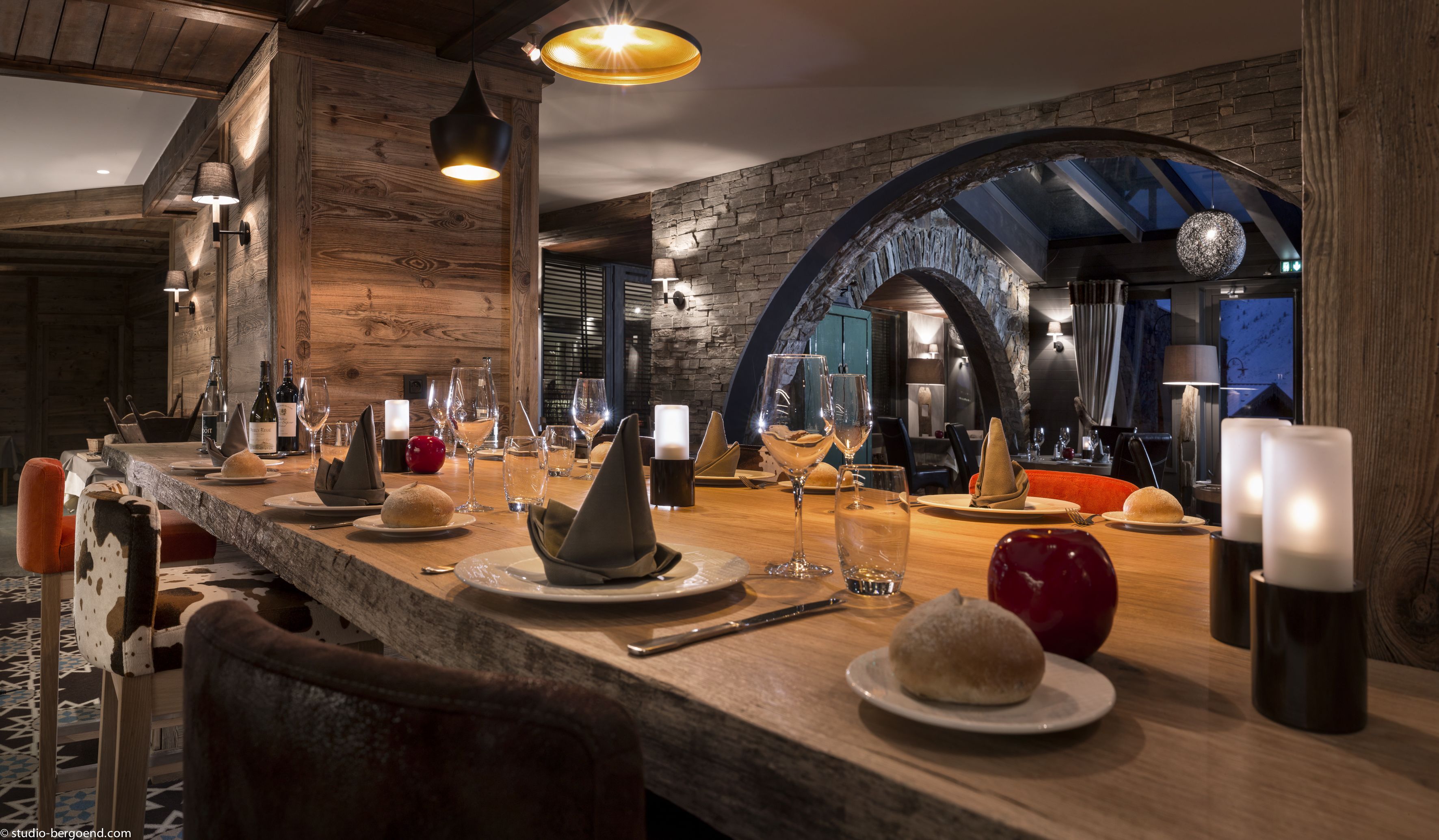 Een restaurant met een rustieke inrichting, met houten balken, stenen muren en een lange eettafel gedekt met kaarsen, glazen en brood.