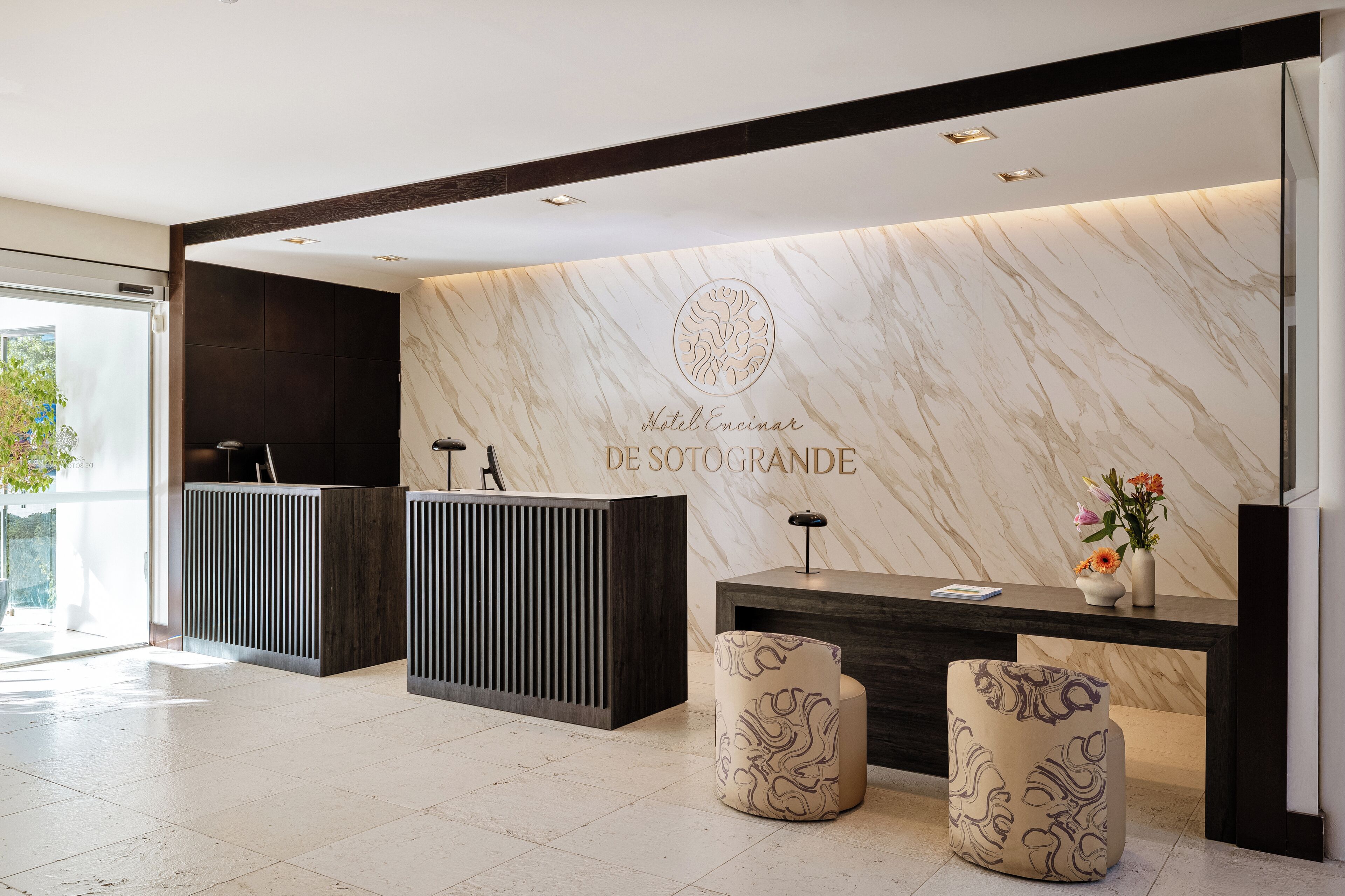 A modern hotel reception area with a marble wall, a reception desk, and two patterned ottomans.