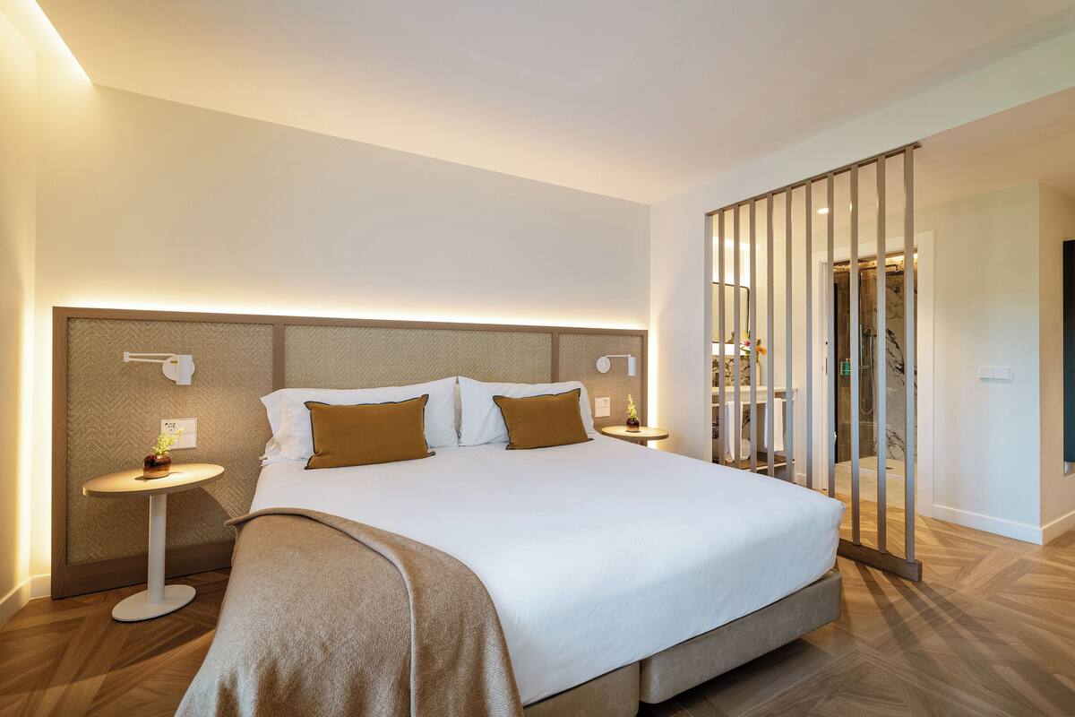A modern hotel room with a large bed, two bedside tables, a headboard with built-in lighting, and a view of the bathroom through a glass partition.