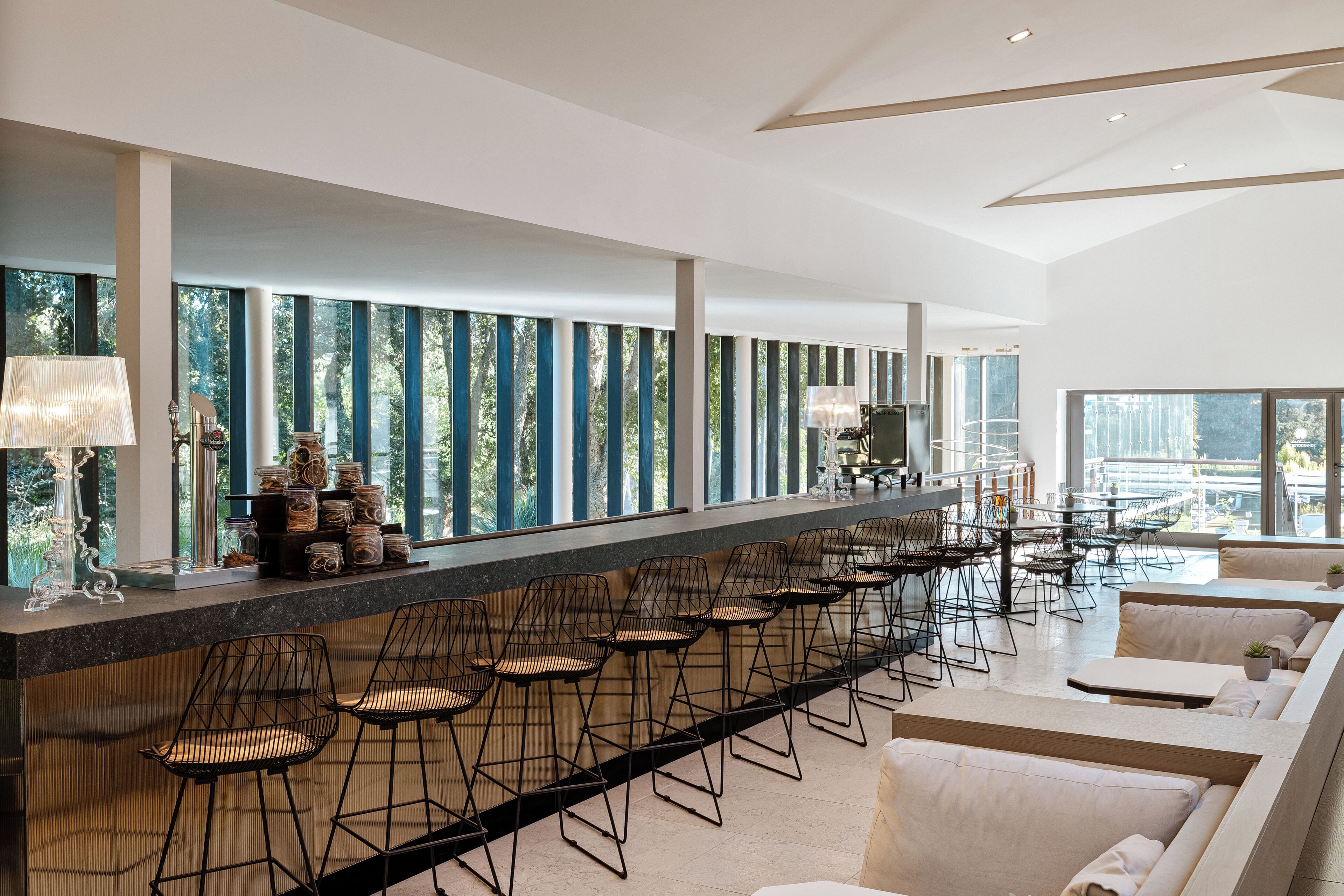 A modern bar area with a long counter, black wireframe chairs, and a seating area with sofas.