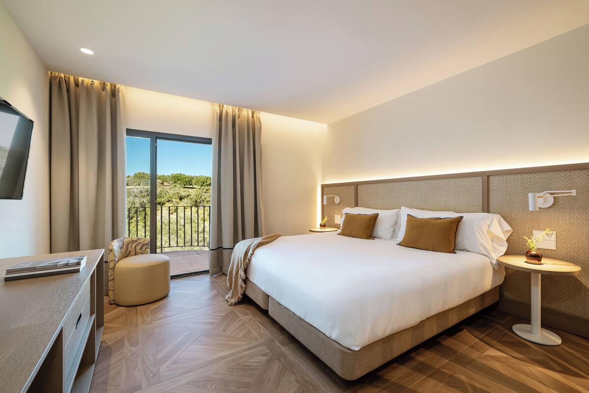 A hotel room with a large bed, a desk, a chair, a small table with a plant, and a balcony with a view of greenery.