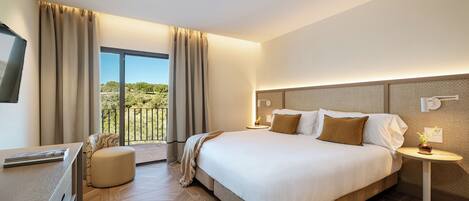 Ett hotellrum med en stor säng, ett skrivbord, en stol, ett litet bord med en växt och en balkong med utsikt över grönområden.