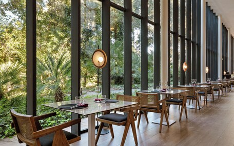 A restaurant with large windows, wooden tables, and chairs, set in a lush green environment.