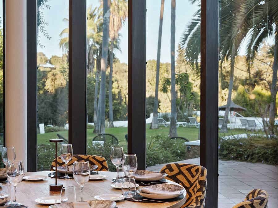 Un comedor con una mesa redonda dispuesta para cuatro personas, ventanas amplias y sillas estampadas.