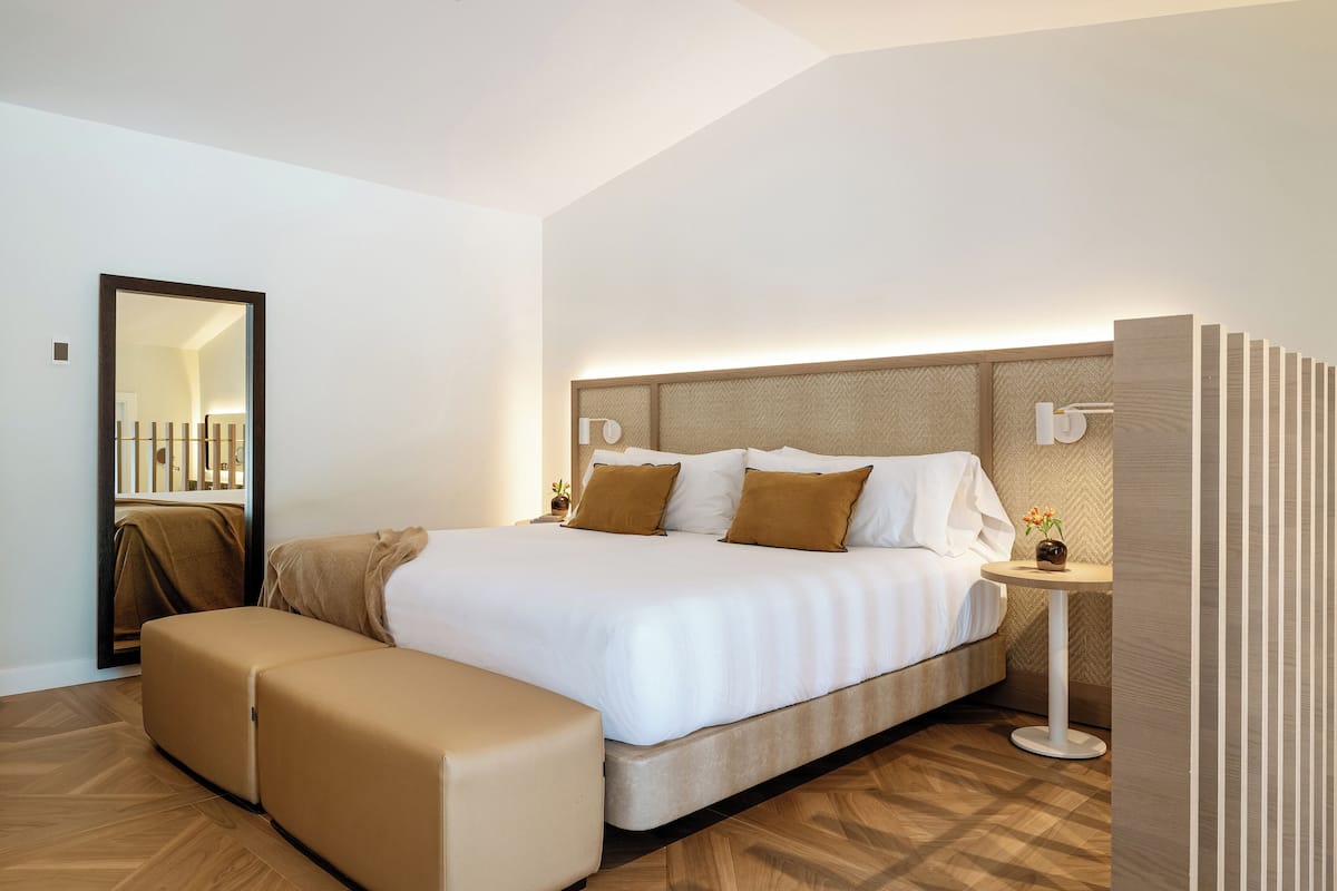 A modern hotel room with a large bed, a bench, a bedside table, and a mirror.