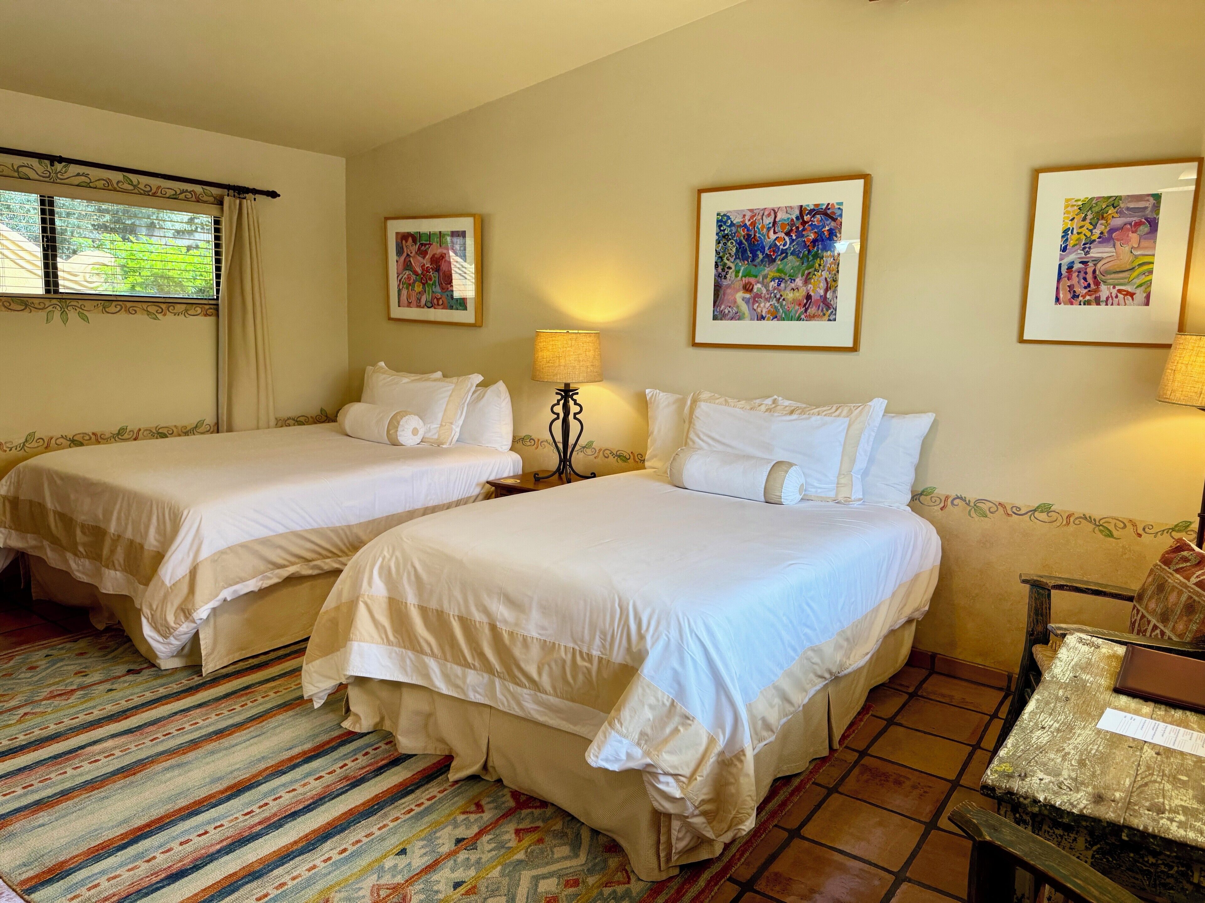 Habitación de hotel con dos camas, una ventana con cortinas, una alfombra estampada y cuadros en las paredes.