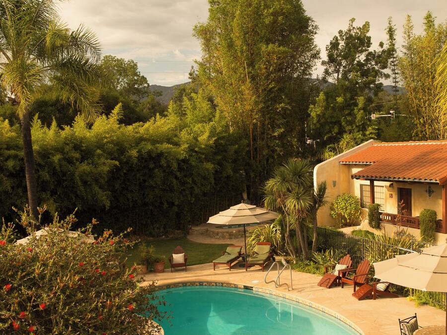 A swimming pool with lounge chairs and umbrellas, surrounded by lush greenery and a house with a red-tiled roof.