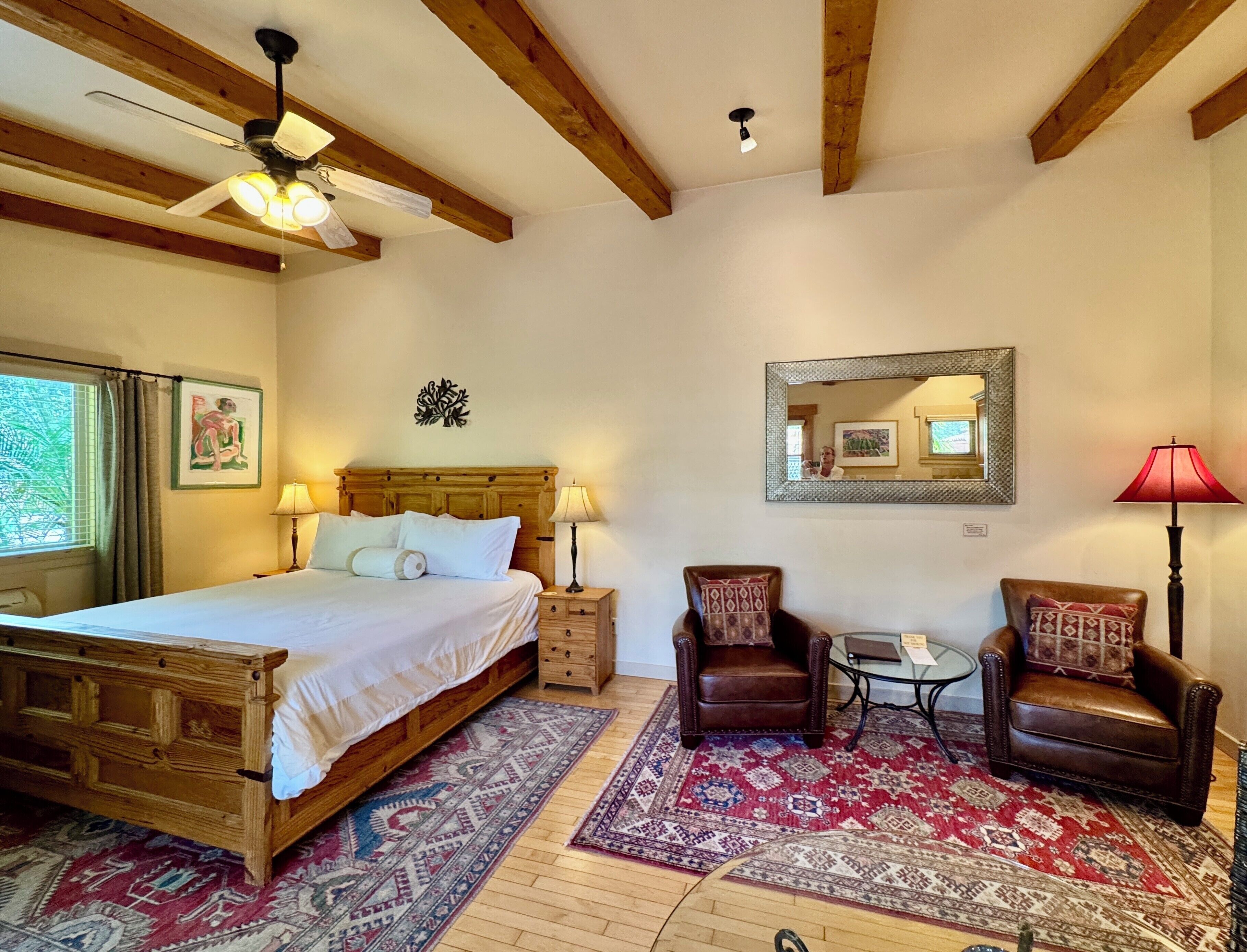 A bedroom with a wooden bed, two chairs, a small table, a mirror, and a ceiling fan.