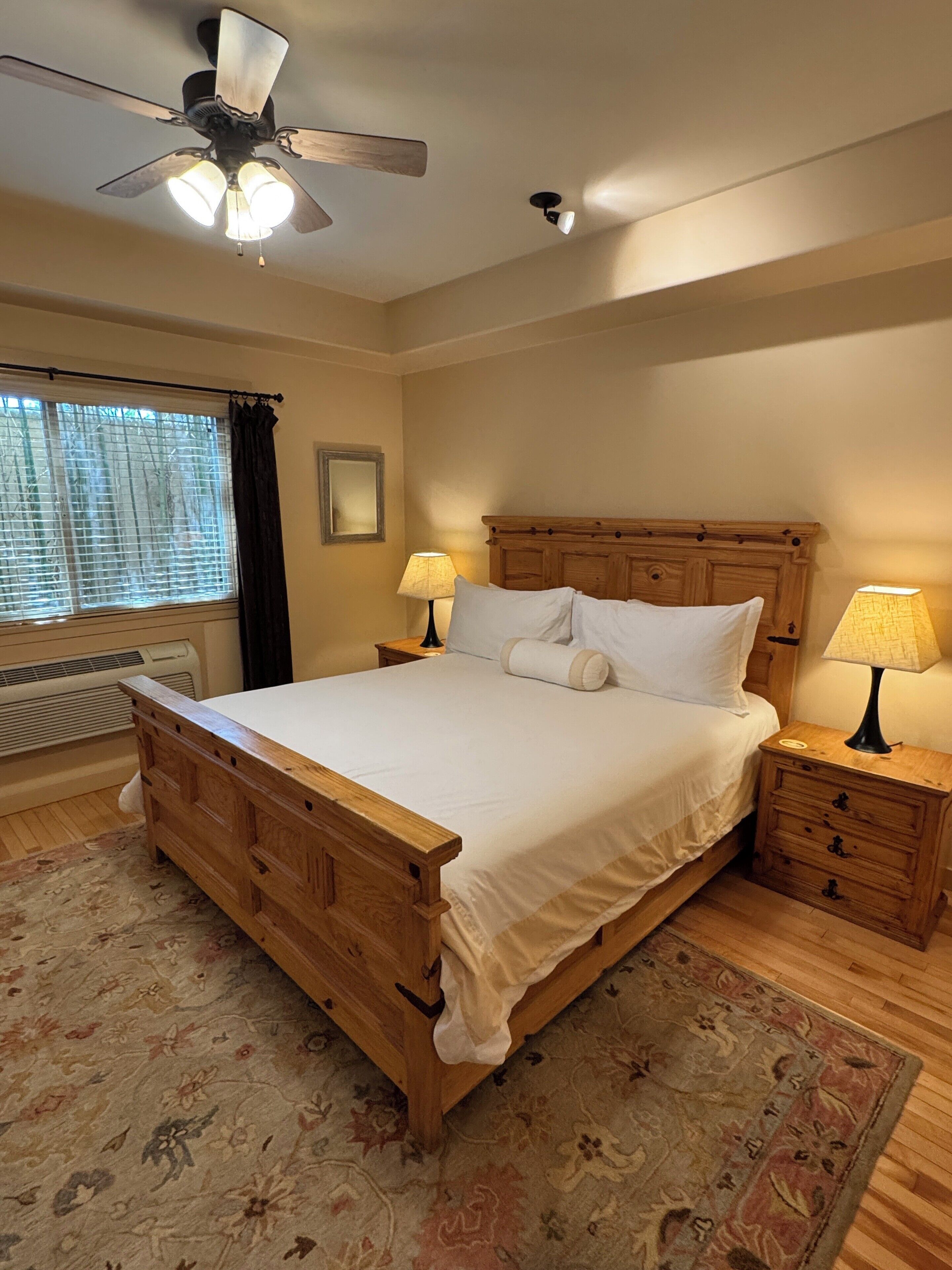 A bedroom with a wooden bed, two bedside tables with lamps, a ceiling fan, a window with blinds, and a radiator.