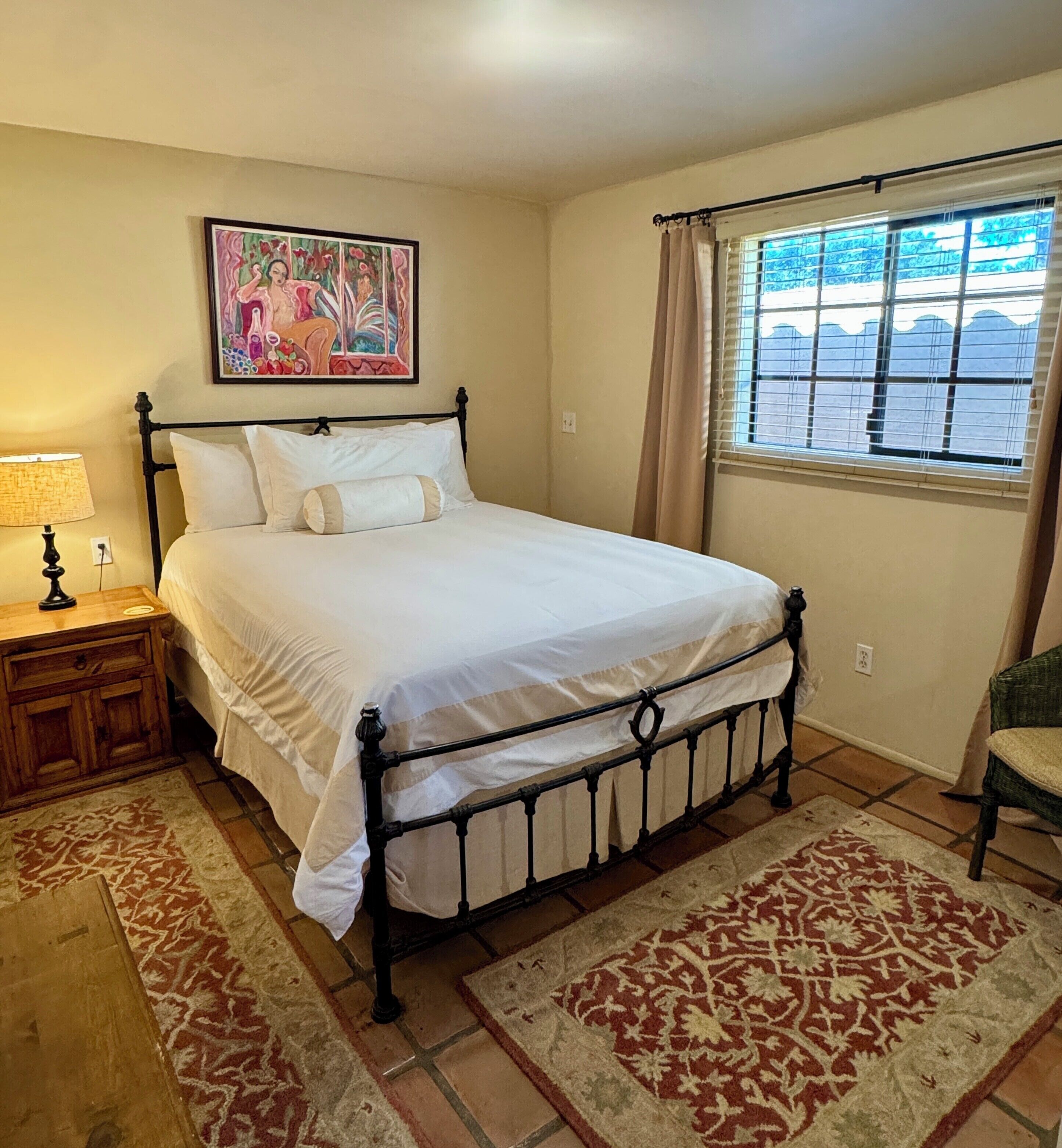 Un dormitorio con cama, mesita de noche, lámpara, ventana con cortinas y un cuadro en la pared.