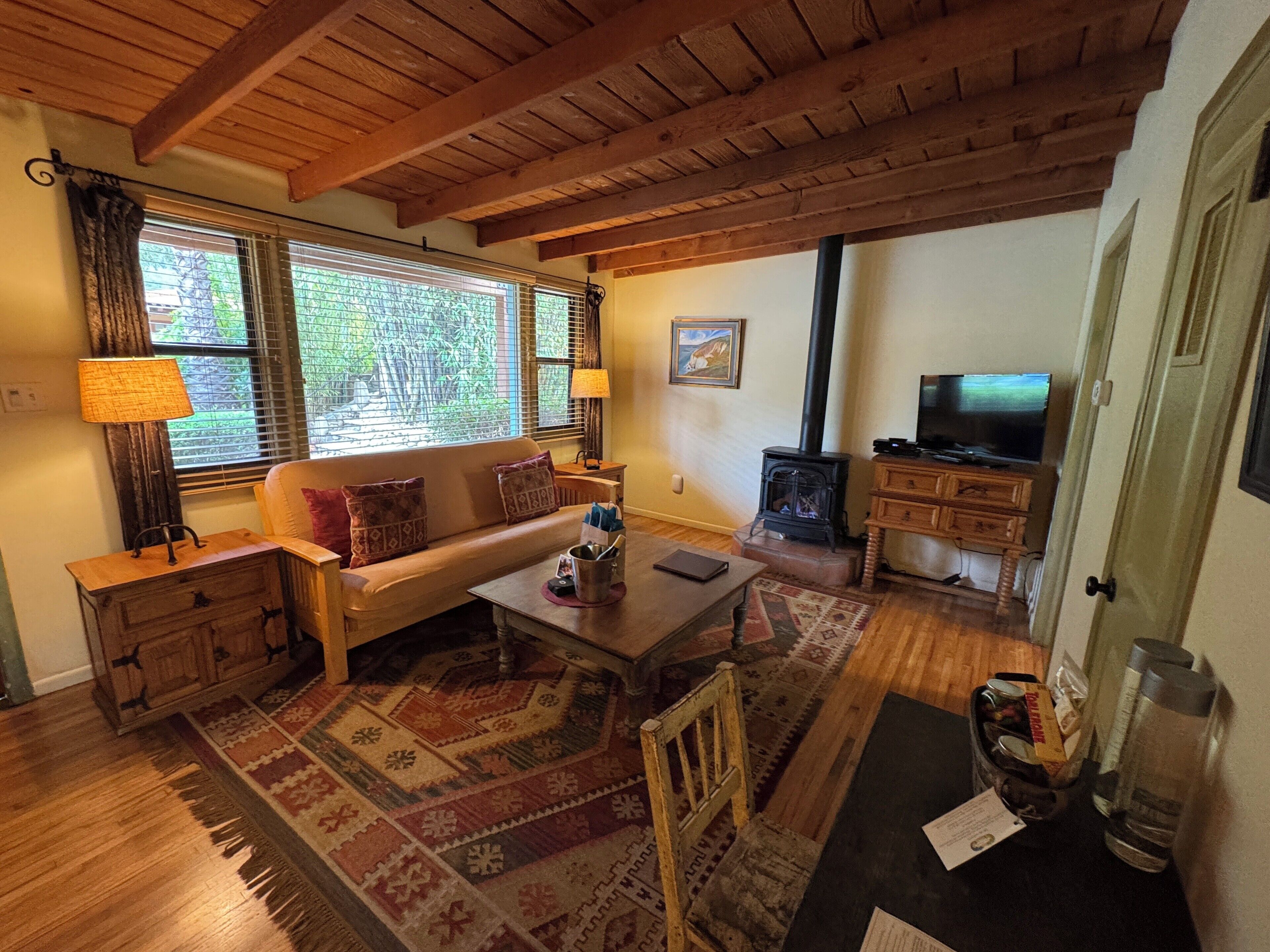 A cozy living room with a sofa, coffee table, and TV. There's a fireplace, wooden beams on the ceiling, and a window with blinds.