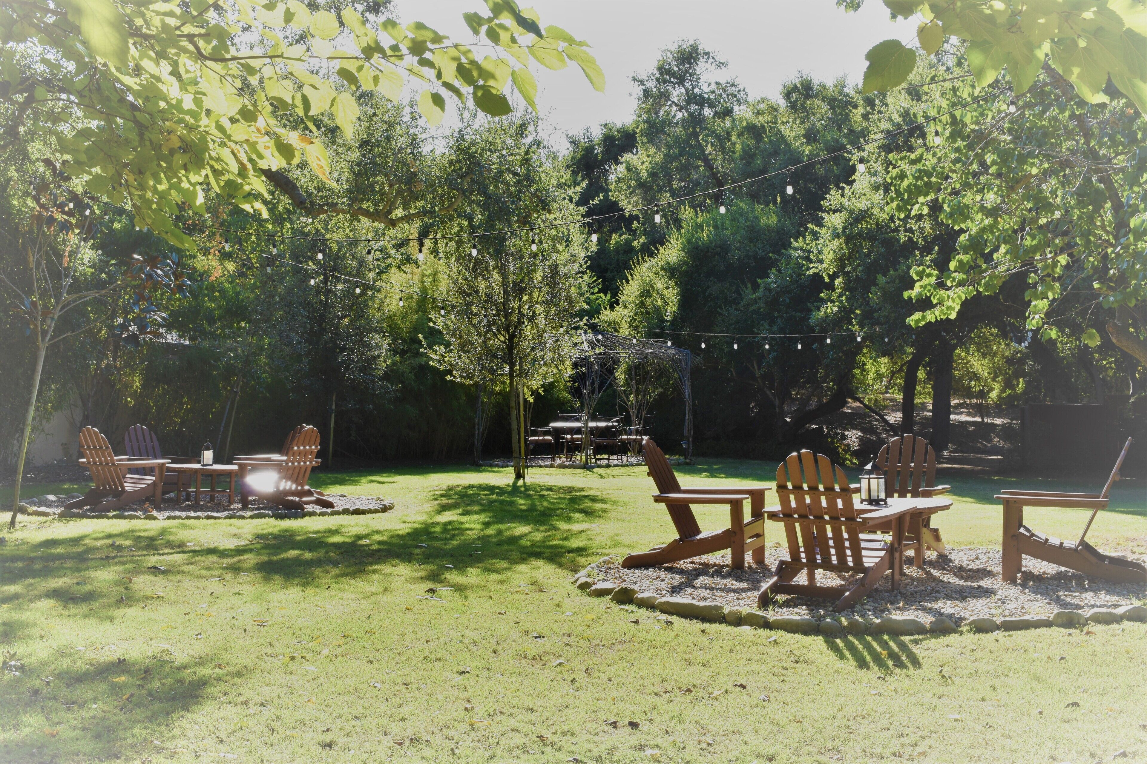 An outdoor seating area with Adirondack chairs and a table, surrounded by trees and a string of lights.