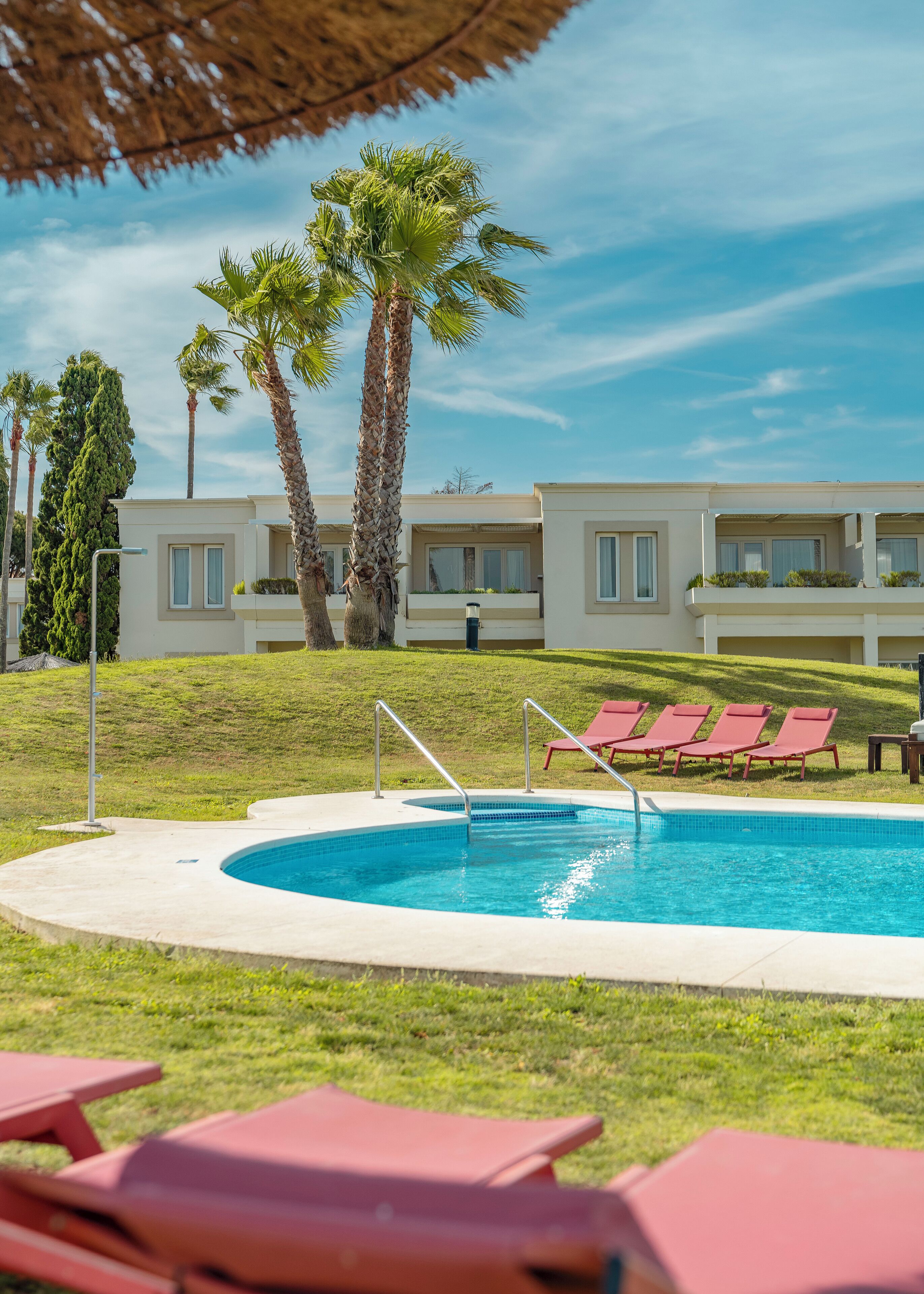 A swimming pool with red lounge chairs, palm trees, and a building with balconies.