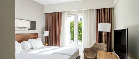 Una habitación de hotel con dos camas, televisión, una silla de mimbre y un ventanal con cortinas.