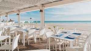 Un restaurante frente al mar con mobiliario de mimbre blanco y mesas dispuestas para comer, con vistas al océano.
