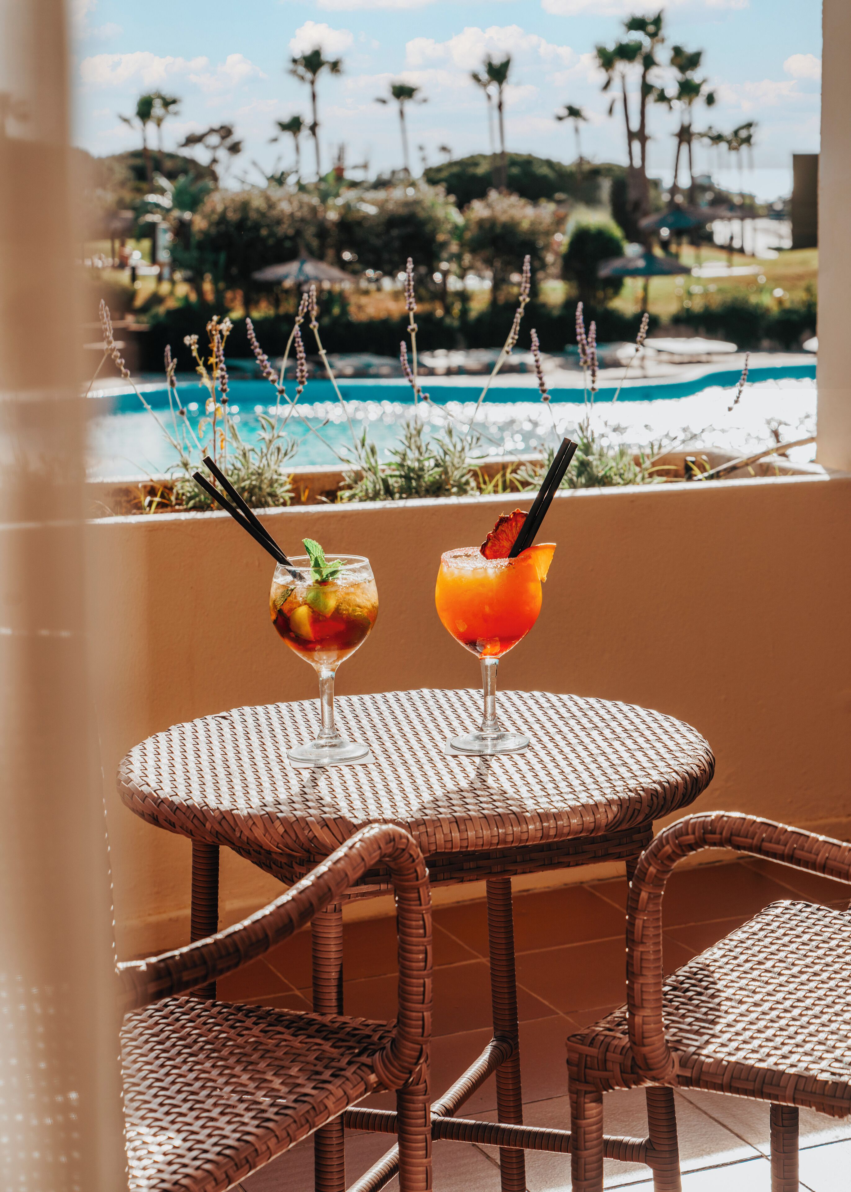 Una mesa de mimbre con dos bebidas, una piscina y palmeras al fondo.