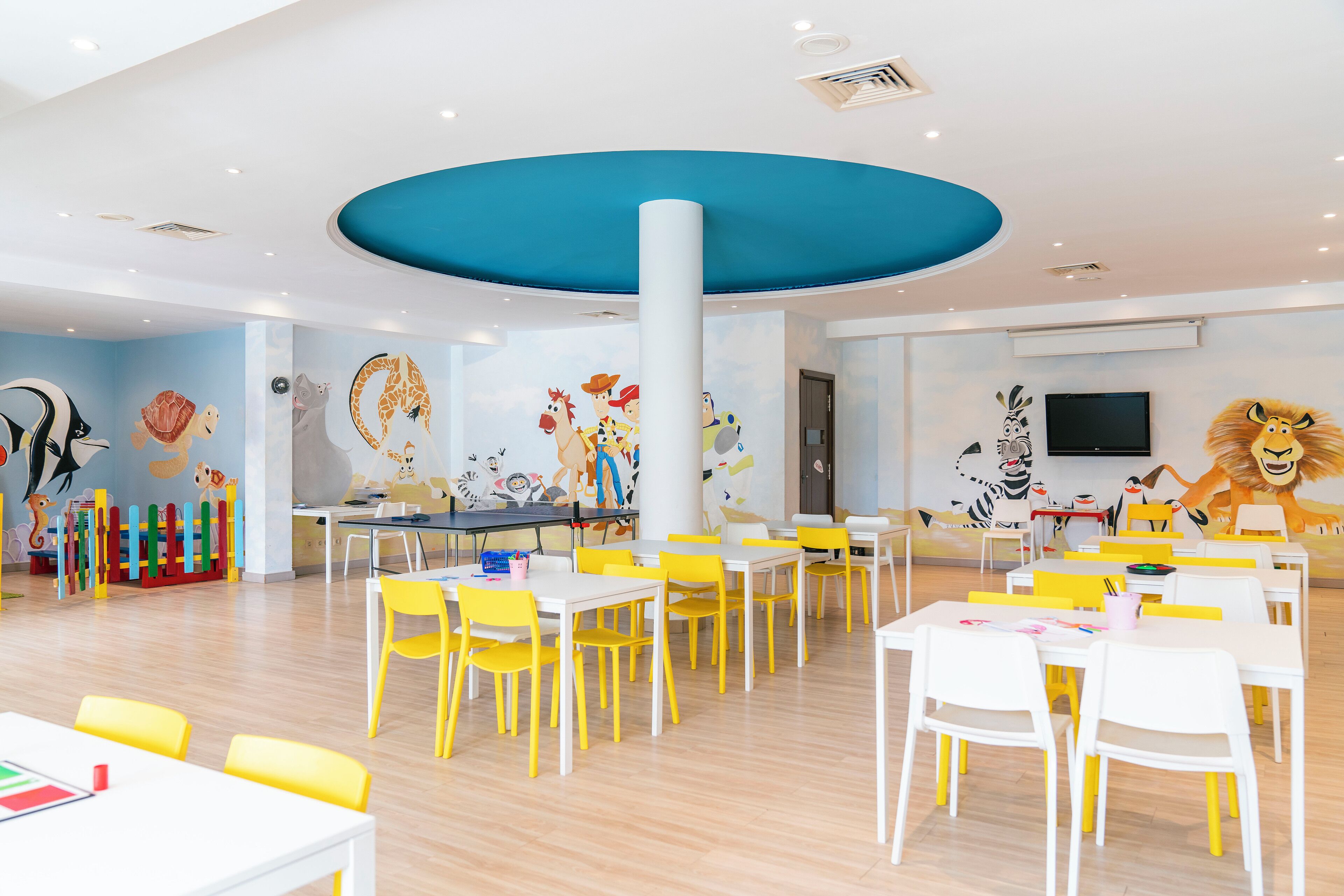Un salón infantil muy luminoso con mesas y sillas amarillas y blancas, un techo azul grande y murales coloridos en las paredes.