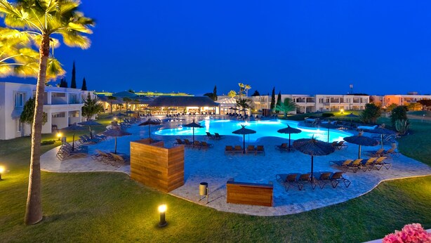 Un resort con una amplia piscina rodeada de tumbonas y sombrillas, un edificio bien iluminado y una palmera.