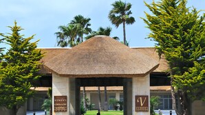 La entrada al Hotel Vincenti Costa Golf, con techo de paja, palmeras y un césped cuidado.