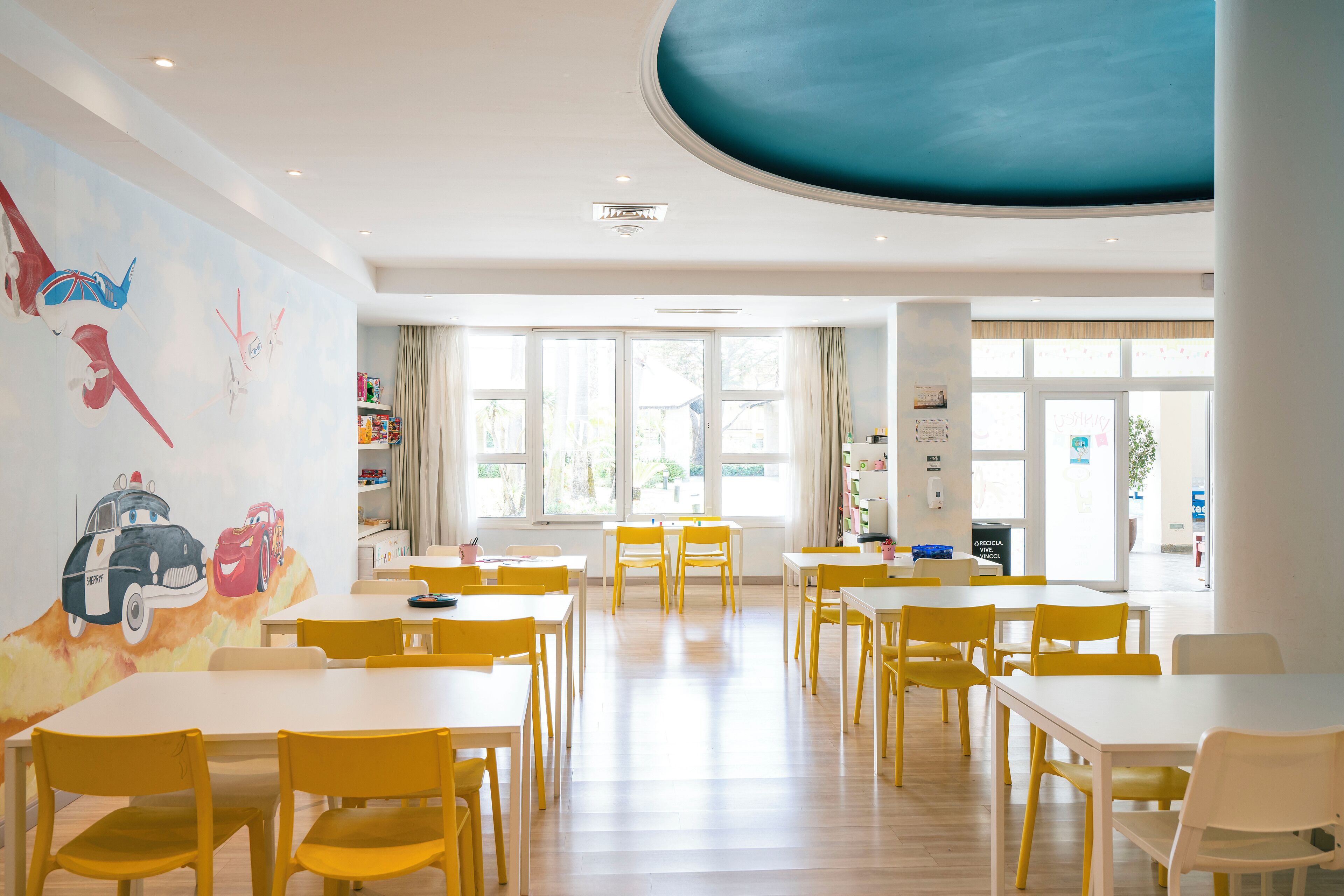 Une pièce lumineuse, avec des chaises jaunes et des tables blanches, de grandes fenêtres et une fresque murale représentant des voitures animées.