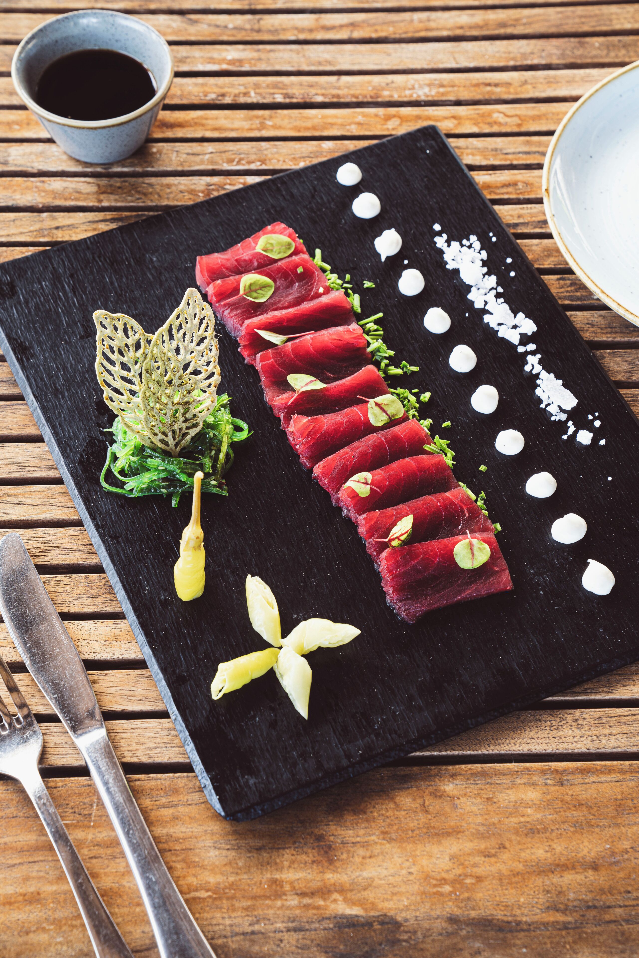 A black slate serving board with a sushi dish, a fork, and a knife on a wooden table.