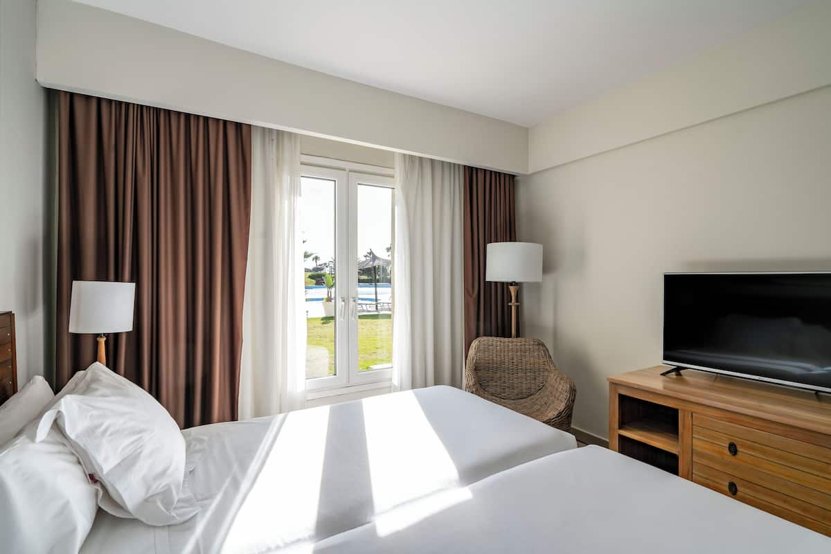 A hotel room with a large bed, two bedside lamps, a television, a wicker chair, and a view of the outdoors through a glass door.