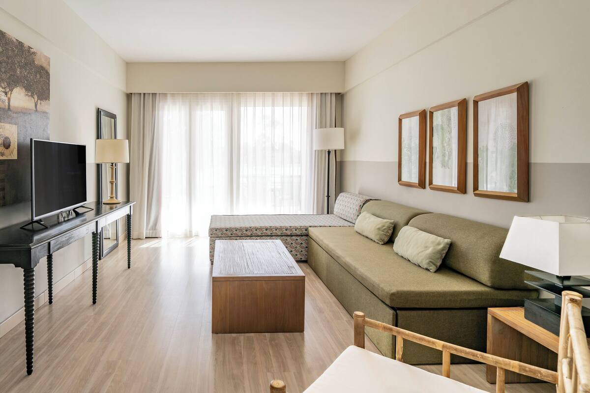 A modern hotel room with a sofa, a TV, a wooden coffee table, and a large window with sheer curtains.