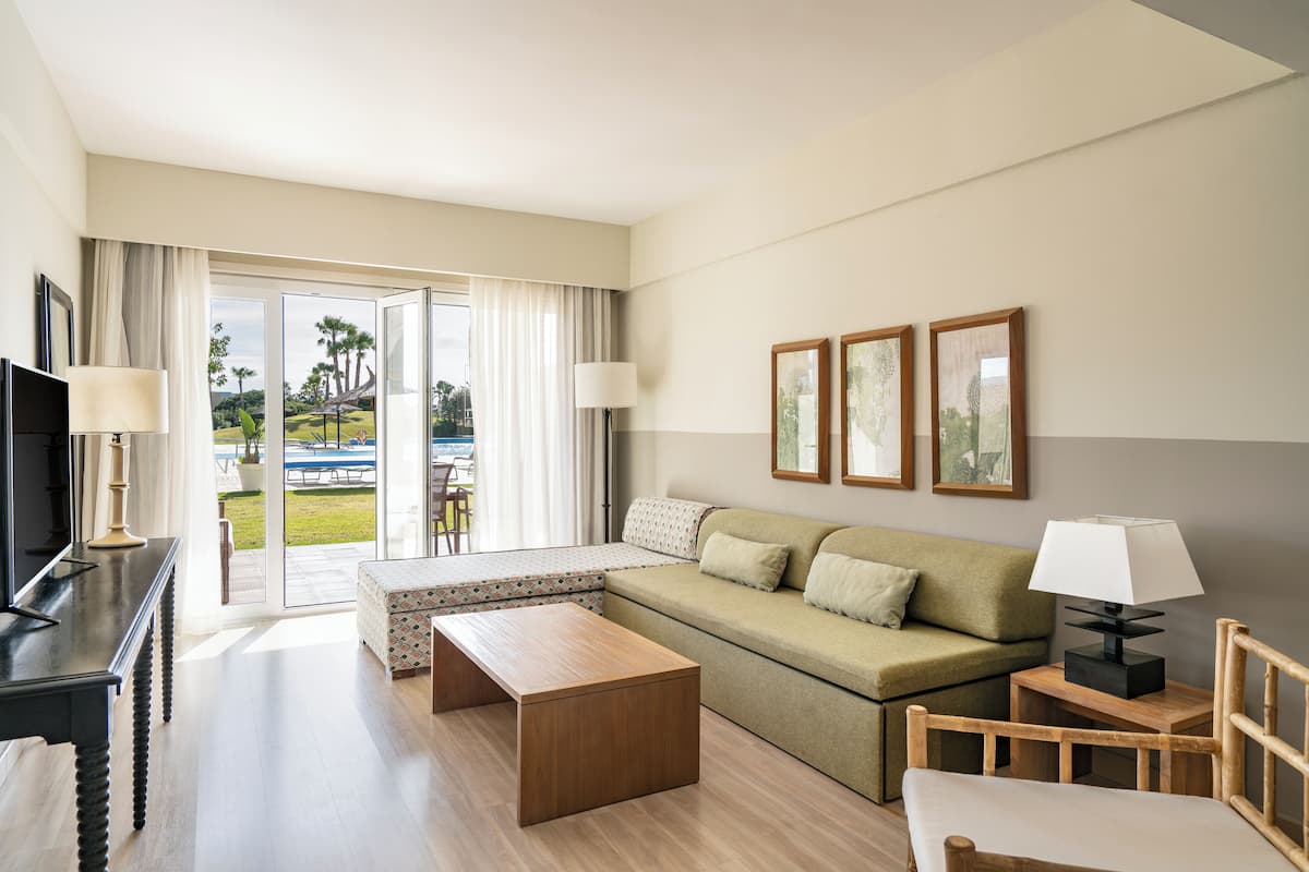 A modern hotel room with a sofa, a wooden coffee table, a TV, and a large window offering a view of the outdoors.