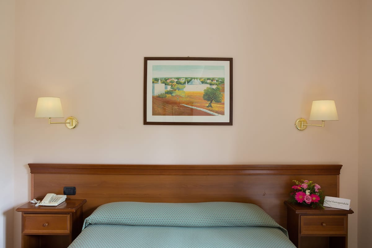 A hotel room with a wooden headboard, a bed with green bedding, two bedside tables with lamps, a telephone, and a framed landscape painting on the wall.