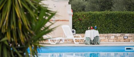 A pool area with lounge chairs and a table with drinks, surrounded by greenery and a stone wall.