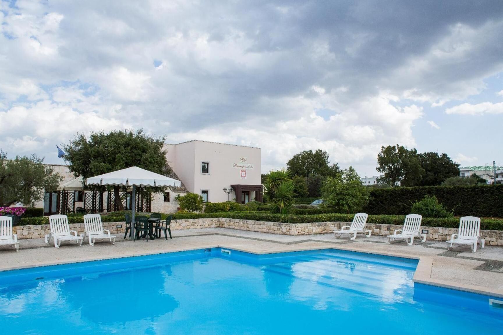 A swimming pool with white lounge chairs and a covered seating area, surrounded by greenery and a building with a sign.