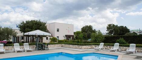 A swimming pool with white lounge chairs and a covered seating area, surrounded by greenery and a building with a sign.