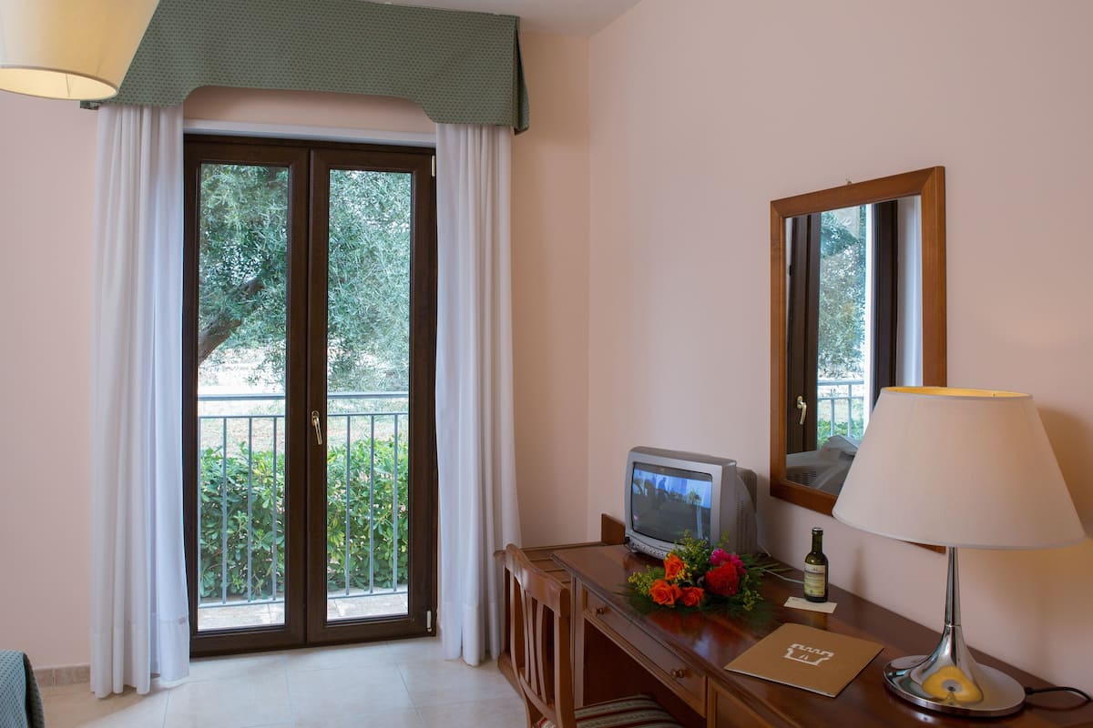 A hotel room with a television on a wooden desk, a mirror, a lamp, and a balcony with a view of greenery.