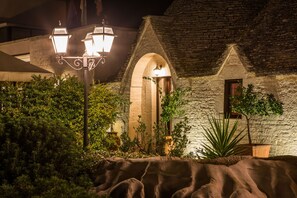 A stone building with an arched doorway and a small window, surrounded by greenery and a rock formation. - Hotel Ramapendula (Alberobello)