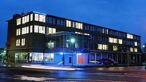 Um hotel moderno com fachada de vidro, vários andares e entrada destacada com toldo iluminado em azul.
