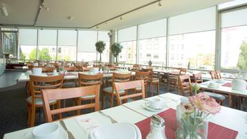 Un comedor amplio con varias mesas y sillas, ventanas grandes con vistas a edificios y árboles, y un jarrón con flores sobre una mesa.