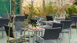Um café ao ar livre com mesas e cadeiras, rodeado por vegetação e edifícios.