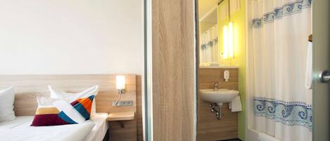 Habitación de hotel con cama, mesita de noche, baño con lavamanos, espejo y cortina de ducha.