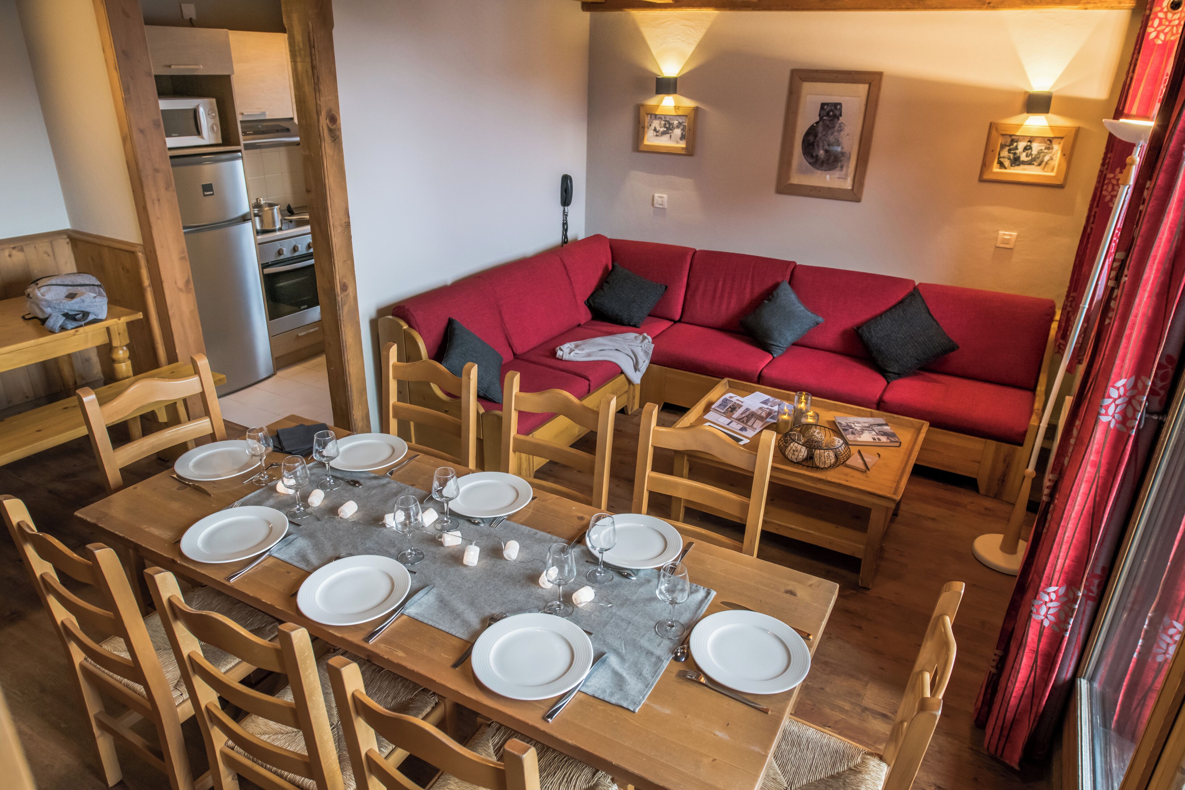 A dining area with a wooden table set for six, a red sofa, wooden chairs, and a kitchen in the background.