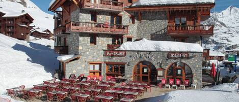 Un edificio de varios pisos con fachada de piedra y balcones de madera, rodeado de terreno cubierto de nieve.