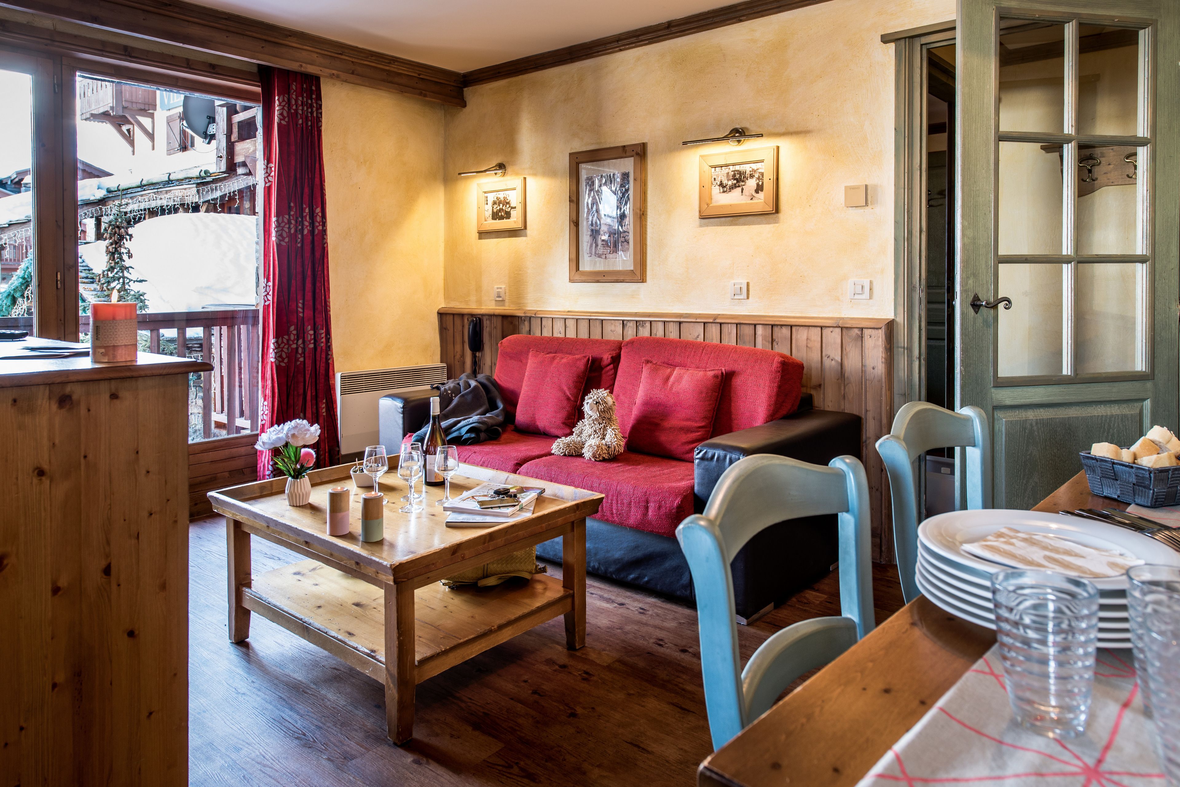 Un salon chaleureux avec une table en bois, un canapé rouge et des fauteuils bleus. Des tableaux encadrés ornent le mur, et une fenêtre offre une vue sur un bâtiment à l’extérieur.