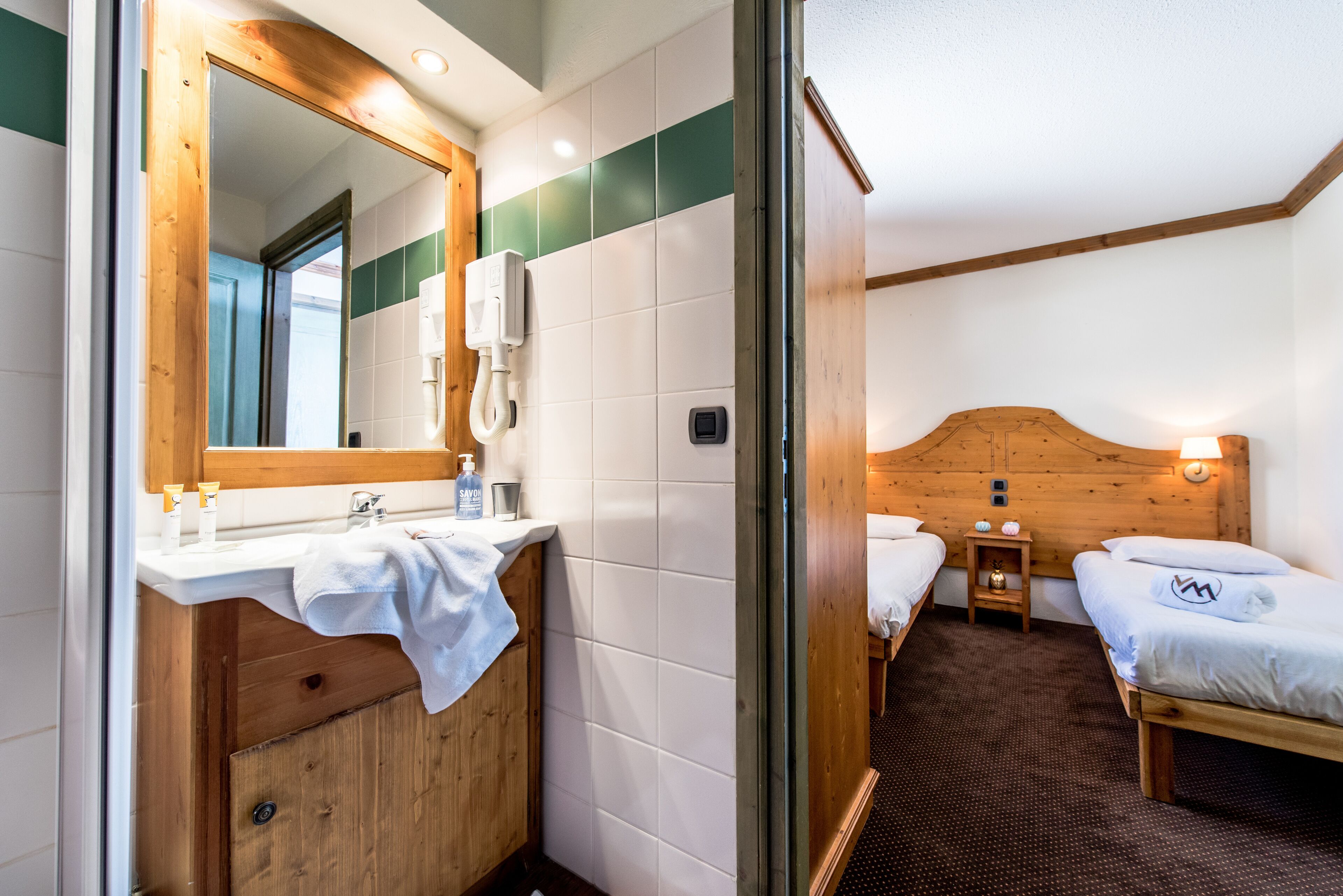 A hotel room with a wooden headboard, a white sink with a mirror, two beds, and a small bedside table.