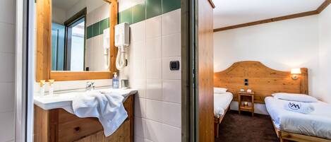 A hotel room with a wooden headboard, a white sink with a mirror, two beds, and a small bedside table.