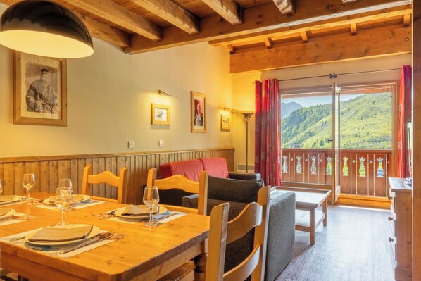 Un comedor con muebles de madera, una mesa dispuesta para dos personas y un balcón con vistas a un paisaje montañoso.