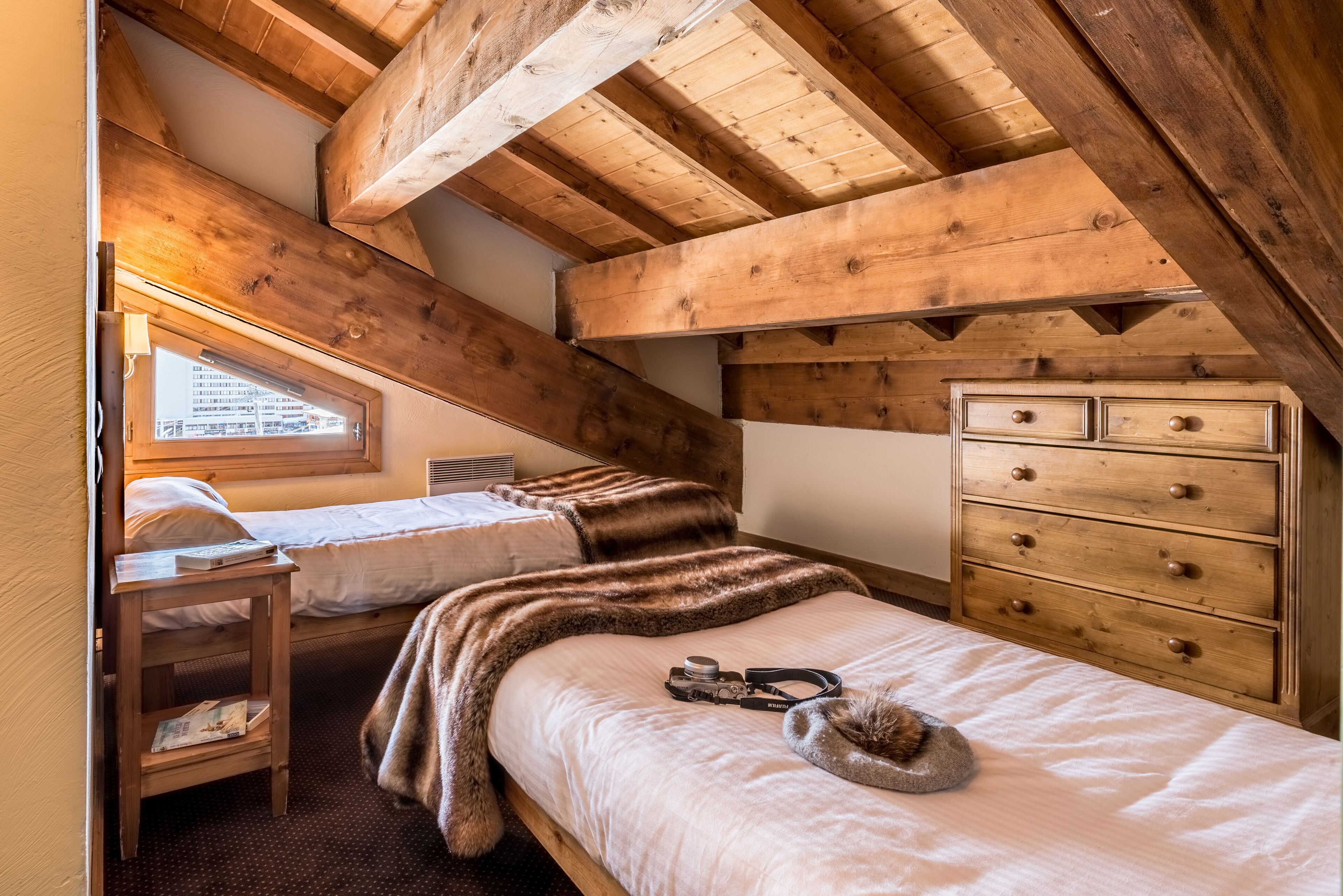A cozy attic bedroom with exposed wooden beams, a bunk bed, a dresser, and a window with a view.