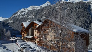 Una cabaña de madera con techo nevado, rodeada de árboles y montañas cubiertas de nieve.